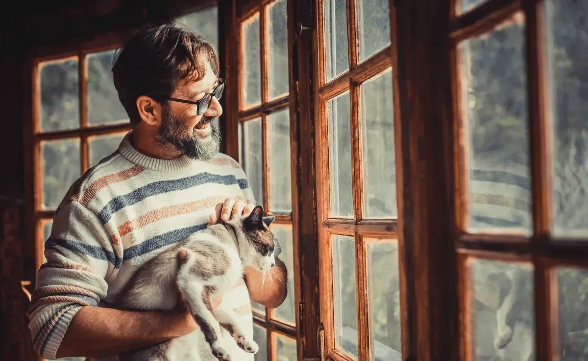 Hombre acariciando a su gato y viendo por la ventana. Foto: Envato/simonapilolla