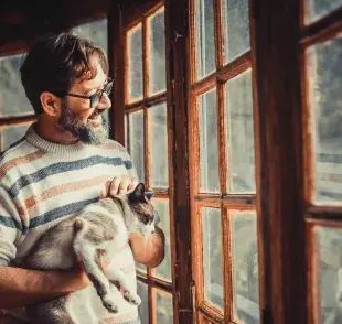 Hombre acariciando a su gato y viendo por la ventana. Foto: Envato/simonapilolla